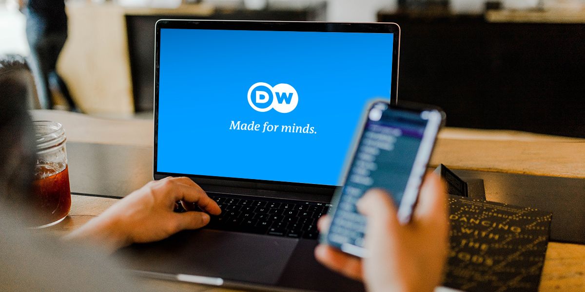 Bild einer Person, die ihr Mobiltelefon in der Hand hält und vor einem Laptop mit dem DW-Logo auf dem Bildschirm sitzt.